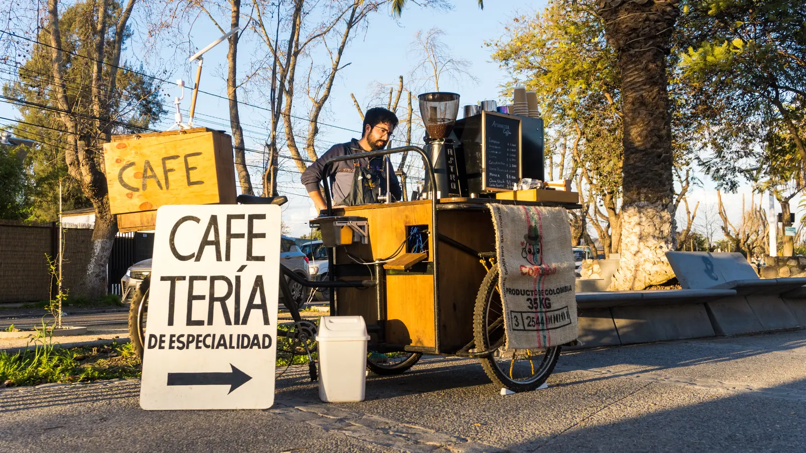 El triciclo cafetería desplegado y funcionando en plena calle, al atardecer, con el barista atendiendo detrás del mesón.